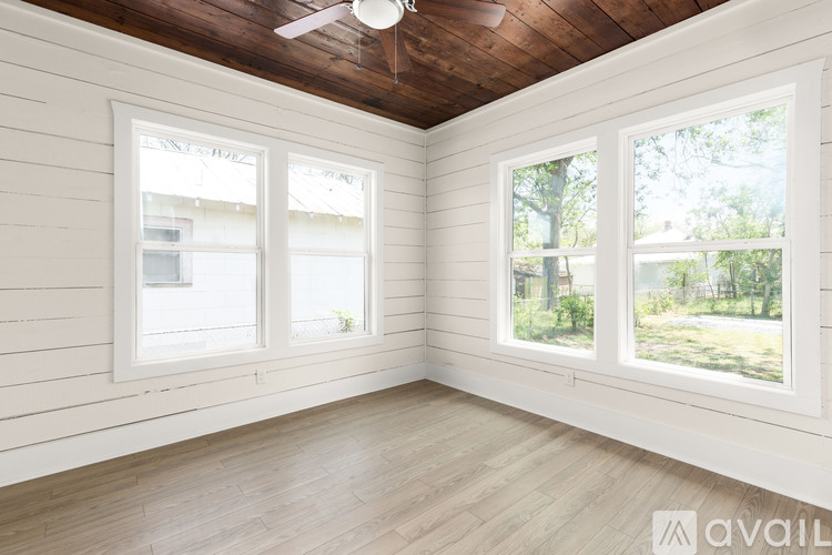 A room with wooden floors and white walls has two windows with a view of trees outside.