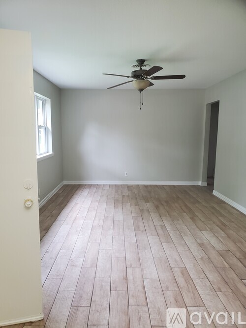 A room with a ceiling fan and light fixture, wooden flooring, and a door on the left.