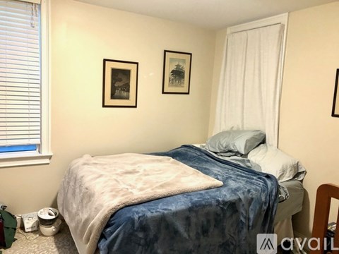 A bedroom with a bed, two pillows, a blanket, a window with blinds, a nightstand with a lamp, a picture on the wall, and a framed picture on the wall.