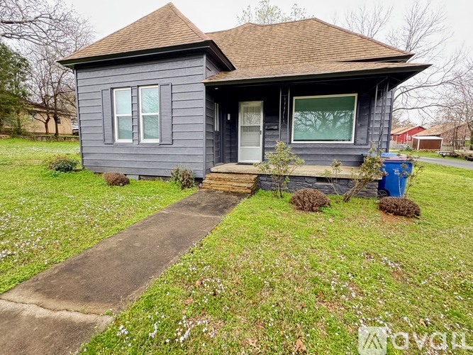 A small house with a brown roof and a grey exterior is surrounded by a grassy area with a few bushes and a blue trash can.