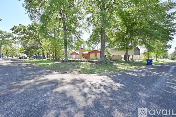 A sunny day at a park with a paved area, trees, and a few buildings in the background.