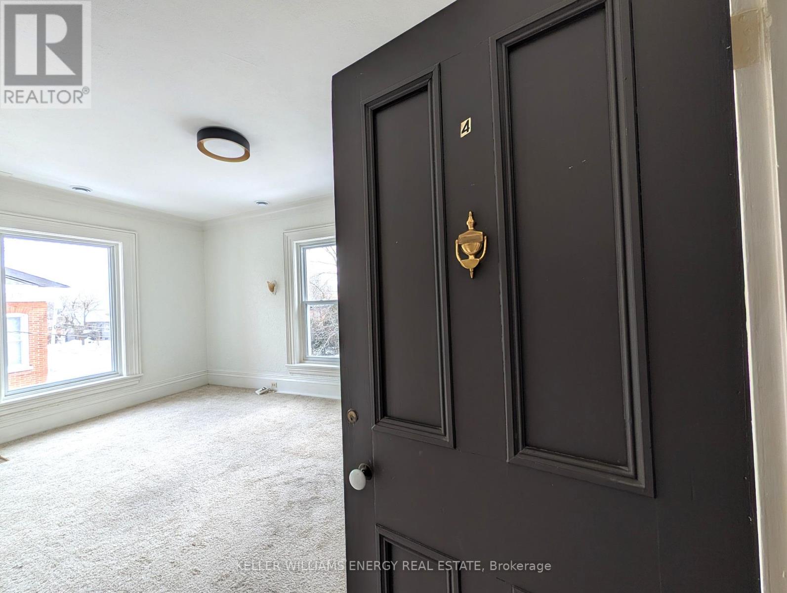 A black door with a brass handle is in the foreground of a room with a carpeted floor and a window.