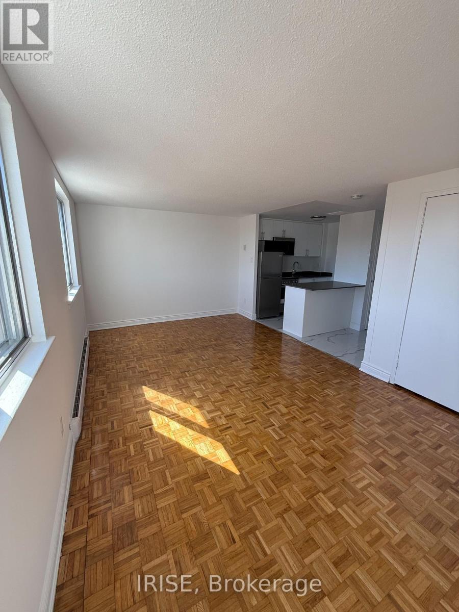 A room with wooden flooring and white walls, with a kitchen area visible in the background.