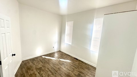 A room with wooden flooring and white walls, with a door on the left and a window on the right.