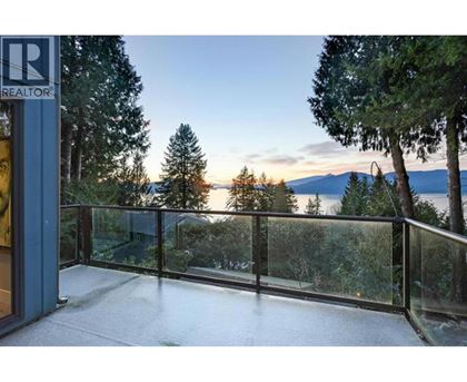 A balcony with a glass railing overlooks a forest and a body of water.