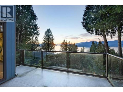 A balcony with a glass railing overlooks a forest and a body of water.