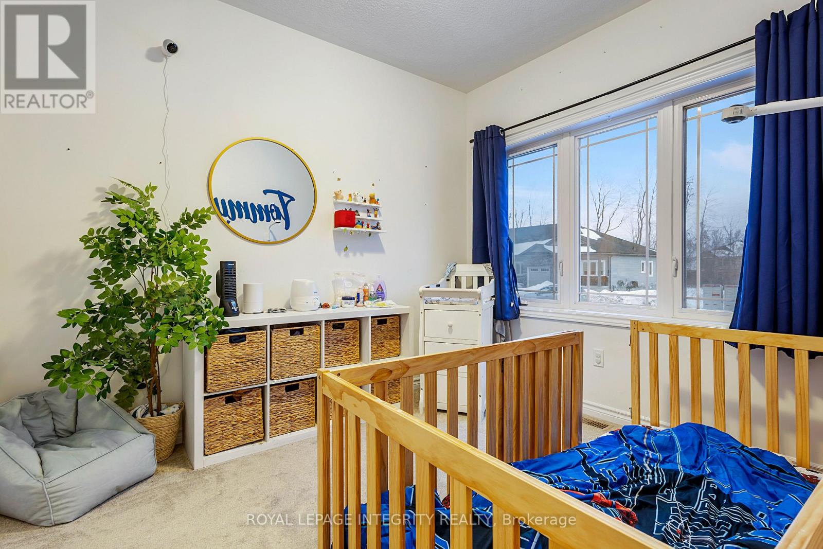 A bedroom with a bed, a window with blue curtains, and a white wall with a round mirror and a logo.
