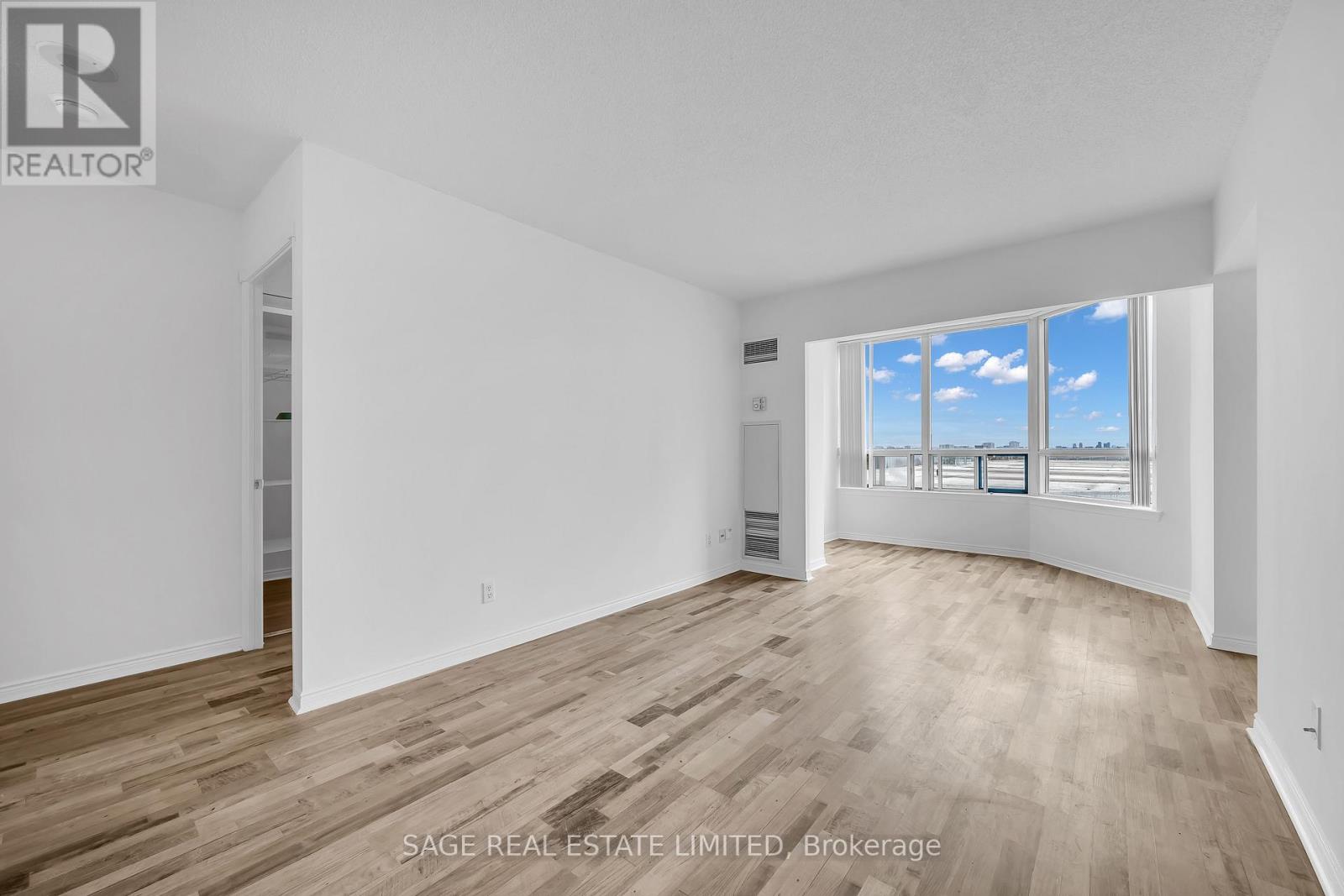 A room with white walls and wooden flooring, with a window showing a blue sky with clouds.