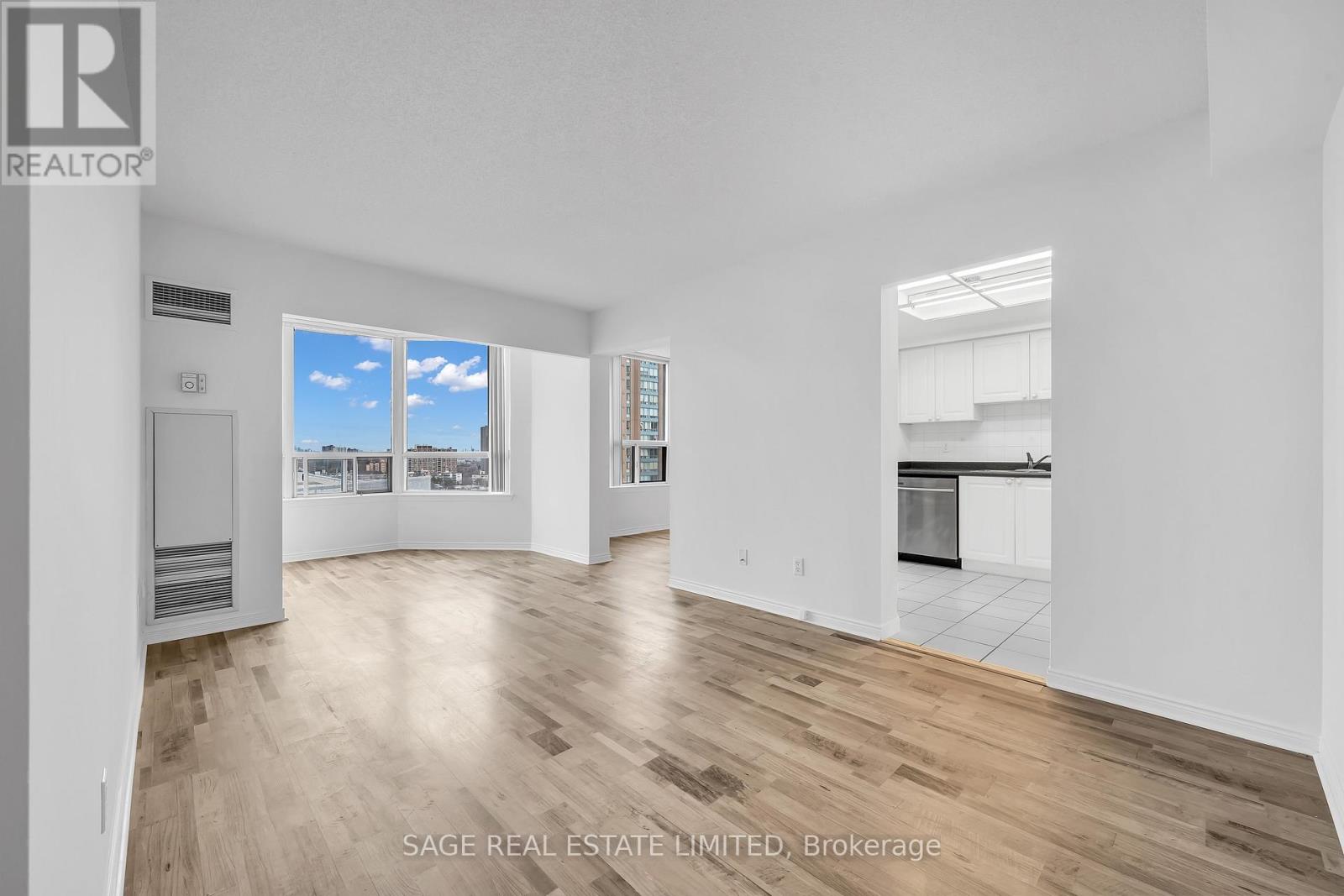 A real estate listing image of a large empty room with wood flooring and a view of the city through the window.