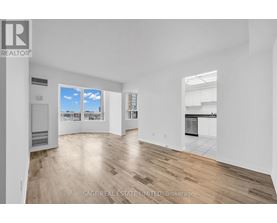 A real estate listing image of a large empty room with wood flooring and a view of the city through the window.