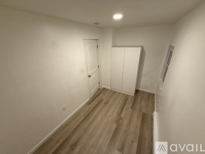 A room with wooden flooring and white walls, featuring a door, a cabinet, and a window.