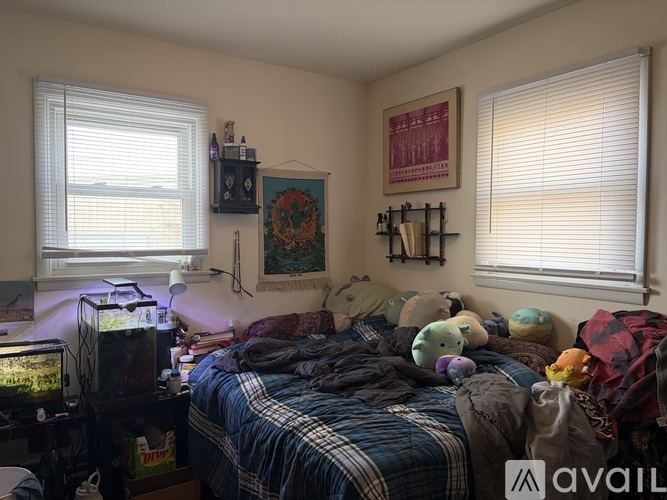 A messy bedroom with a bed covered in blankets and stuffed animals, a fish tank on a table, and a wall hanging.