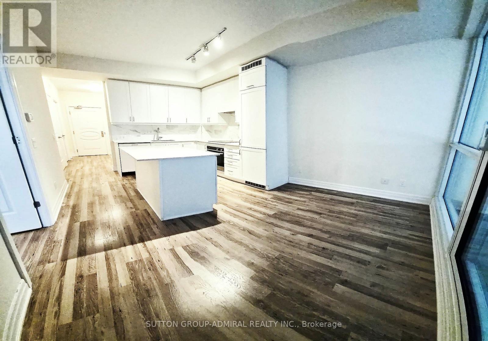 A room with wooden floors and white walls, with a kitchen area in the background.