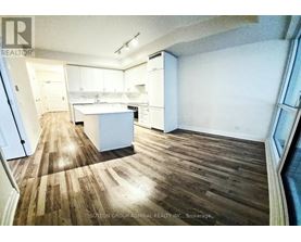 A room with wooden floors and white walls, with a kitchen area in the background.