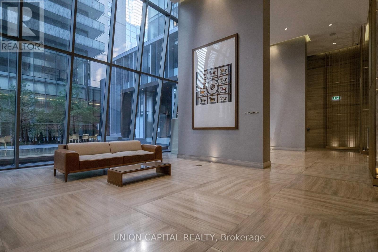 A large artwork hangs on a wall in a spacious room with a wooden floor and a glass wall.