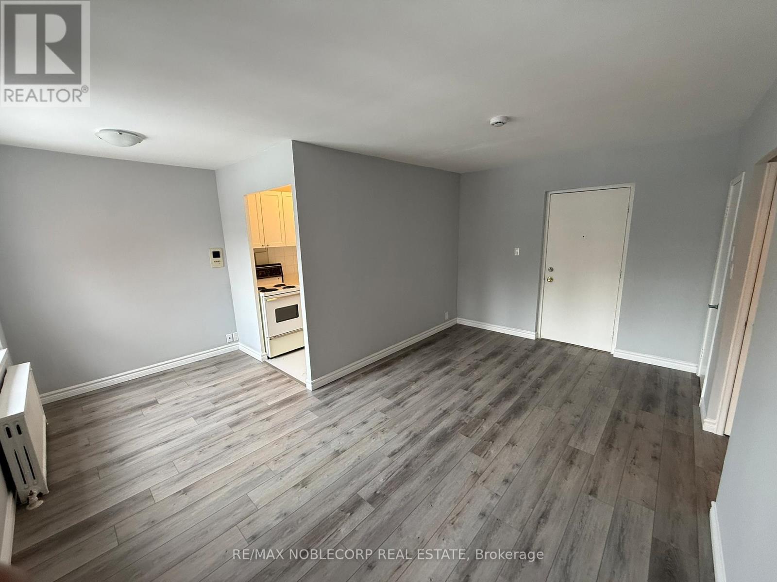 A room with grey walls and wooden flooring, with a white door and a small kitchenette visible.