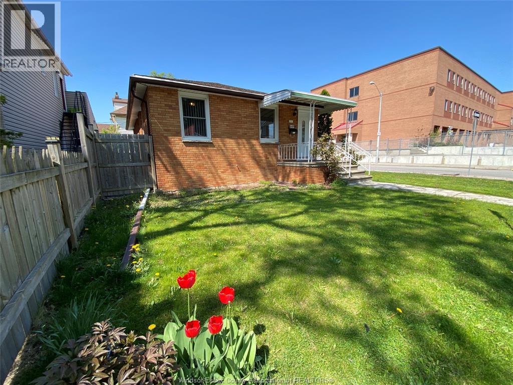 A small red brick house with a white door and windows, a wooden fence, and a green lawn with red tulips.