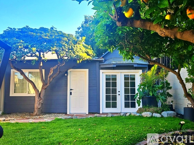 A blue house with a white door and windows, surrounded by greenery and trees.