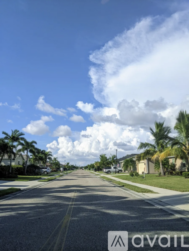 A street with palm trees and houses on both sides under a blue sky with clouds.