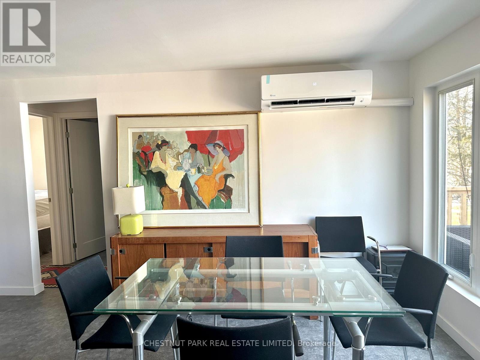 A dining room with a glass table and chairs, a painting on the wall, and a window with a view of trees outside.