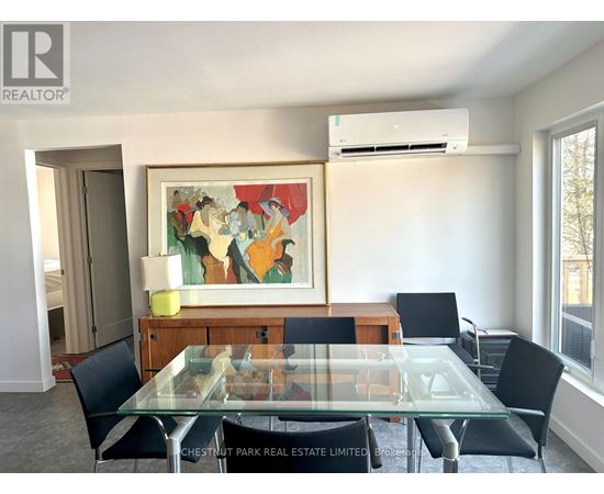 A dining room with a glass table and chairs, a painting on the wall, and a window with a view of trees outside.