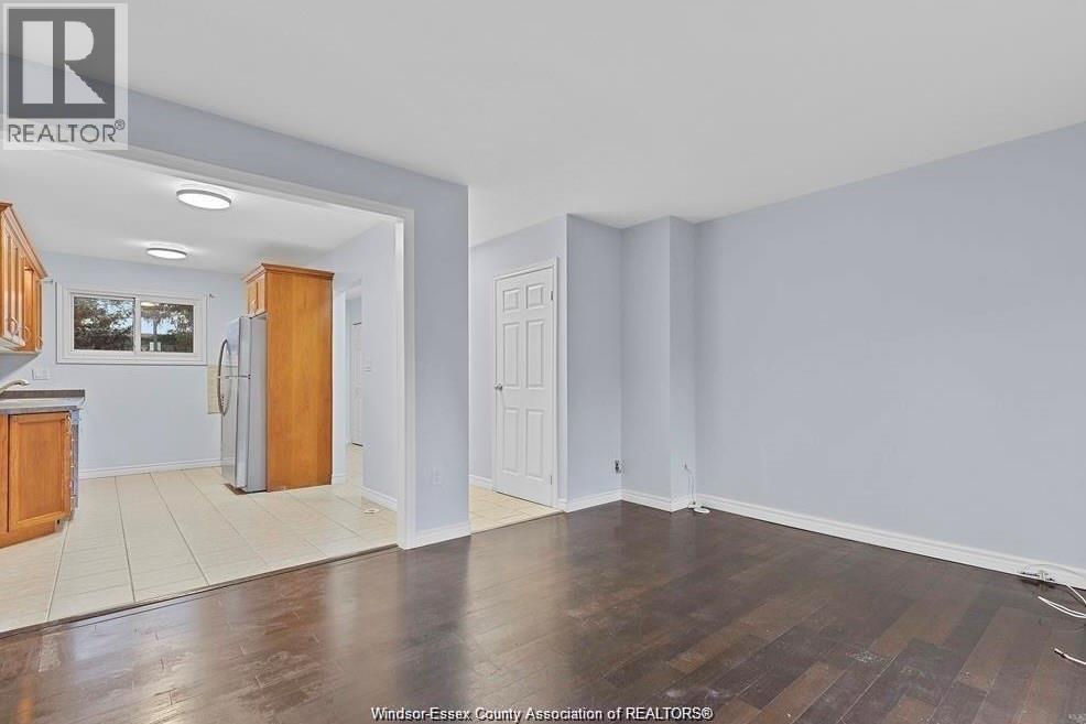 A room with wooden floors and white walls, with a kitchen area visible in the background.