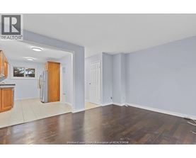 A room with wooden floors and white walls, with a kitchen area visible in the background.