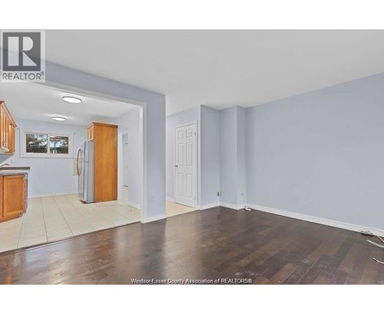 A room with wooden floors and white walls, with a kitchen area visible in the background.