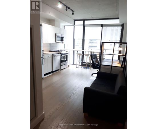 A living room with a black couch and a kitchen area with white appliances.