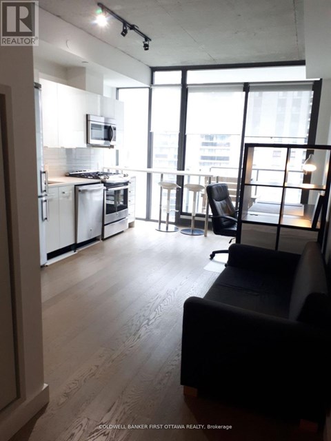 A living room with a black couch and a kitchen area with white appliances.