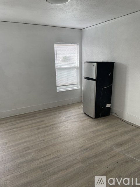 A black fridge sits in a room with a window and wooden flooring.