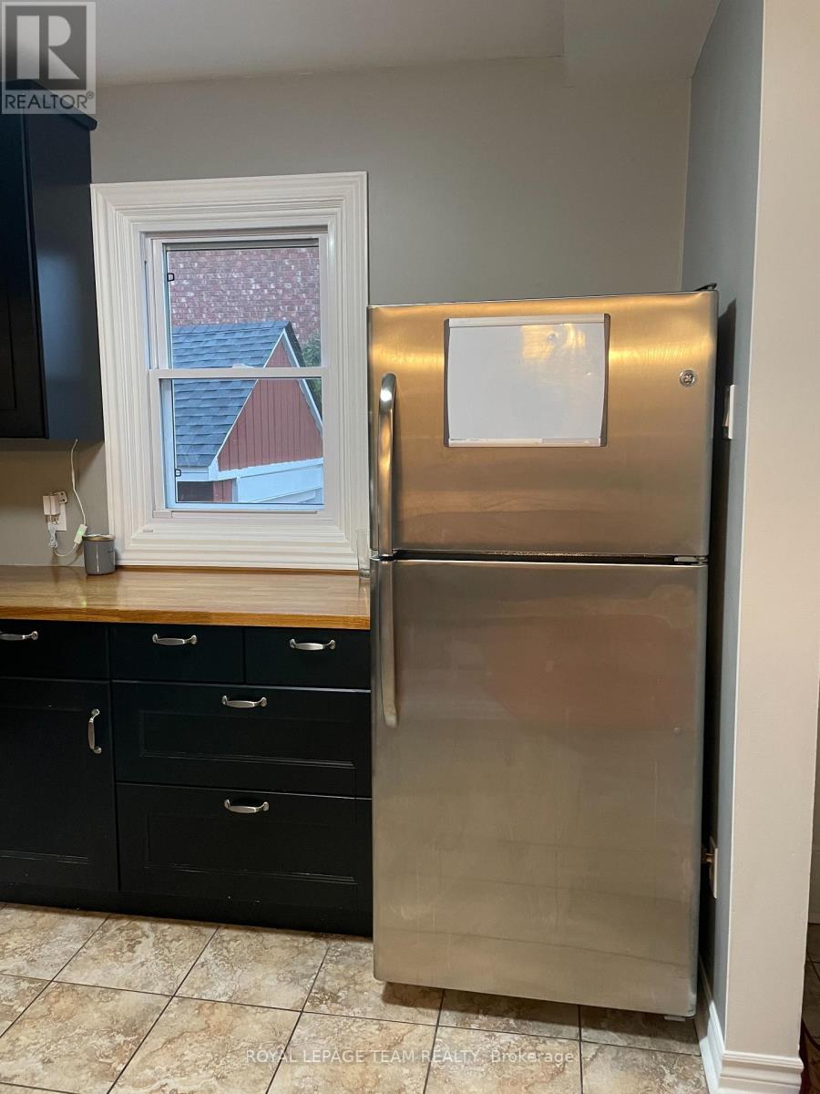 A stainless steel refrigerator stands in a kitchen with black cabinets and a window showing a red house in the background.