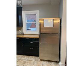 A stainless steel refrigerator stands in a kitchen with black cabinets and a window showing a red house in the background.