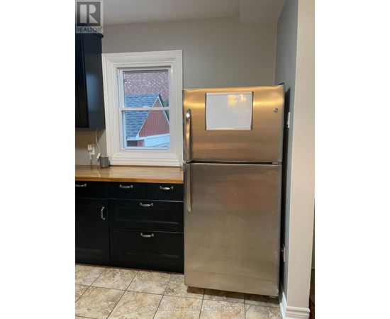 A stainless steel refrigerator stands in a kitchen with black cabinets and a window showing a red house in the background.