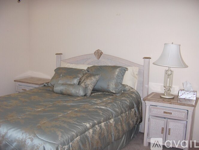 A bed with a grey comforter and two grey pillows sits next to a nightstand with a white lamp and tissue box.