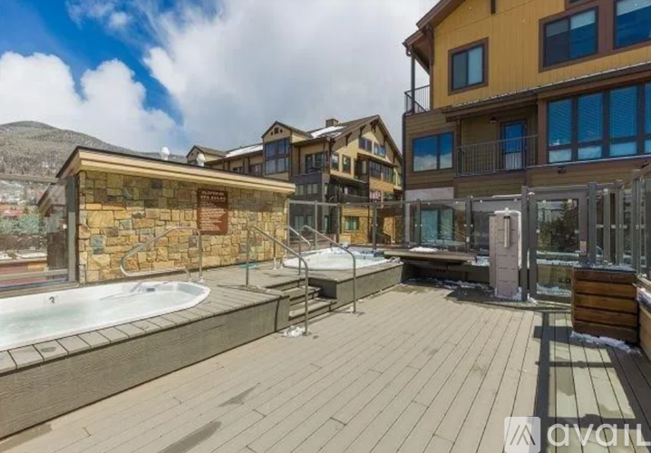 A hot tub sits on a wooden deck in front of a stone wall and a building.