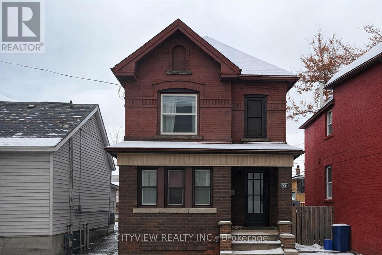 A red brick house with a black door and windows is in front of a grey house and a red building.