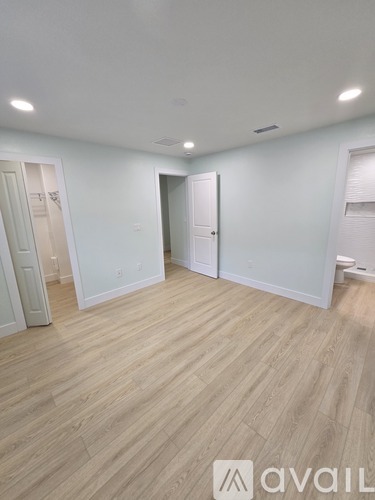 A room with light wood flooring and white walls, with a door on the right and a window on the left.