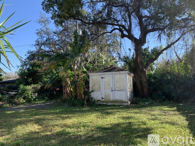 A small white shed sits in a grassy area with trees and bushes around it.