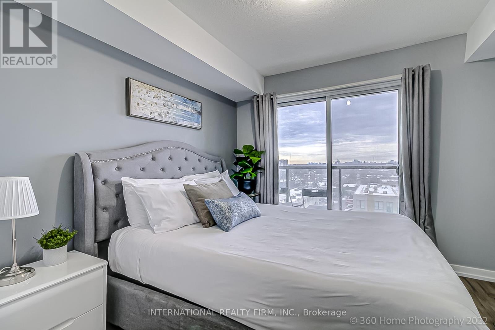 A bedroom with a large bed, a nightstand with a lamp, and a window with a view of the city.