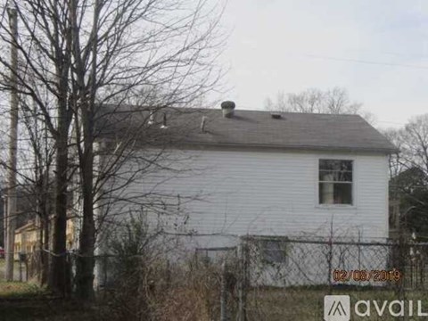 A white house with a grey roof and a chimney is surrounded by a fence and leafless trees.
