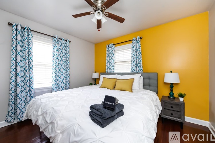 A bedroom with a white bed, blue and white patterned curtains, a ceiling fan, and a small nightstand with a lamp.
