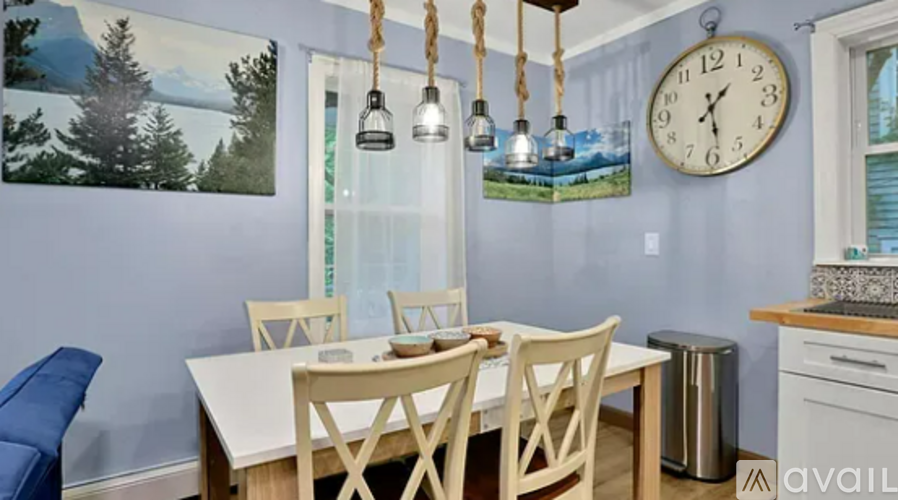 A dining room with a white table and chairs, a clock on the wall, and a painting of a forest scene.