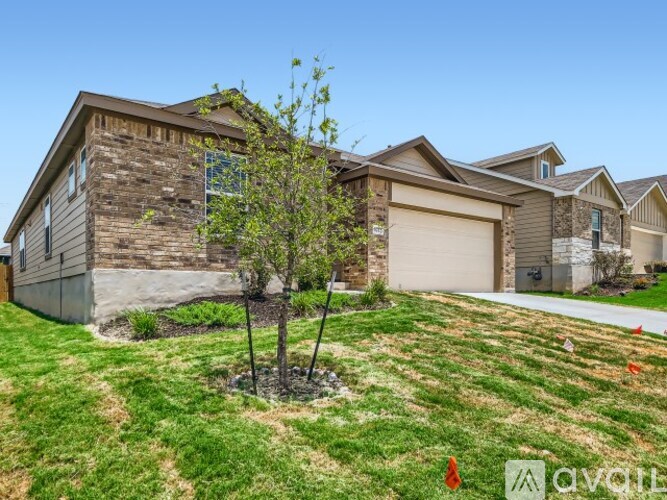 A house with a brown stone facade and a white garage door is surrounded by a grassy lawn with a young tree in the foreground.