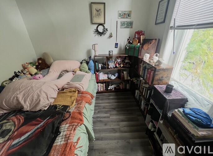 A messy bedroom with a bed covered in blankets and pillows, a bookshelf filled with items, and a window with blinds.