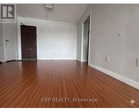 A room with wooden flooring and white walls, with a door on the left and a window on the right.