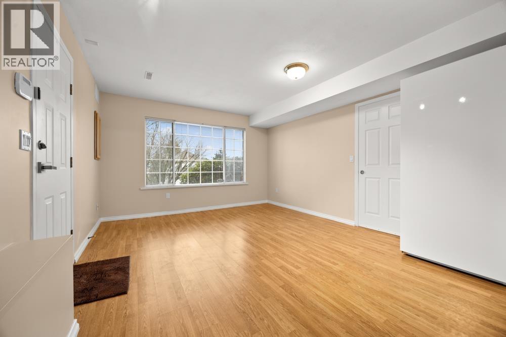 A spacious room with wooden floors and white walls, featuring a large window with a view of trees outside.