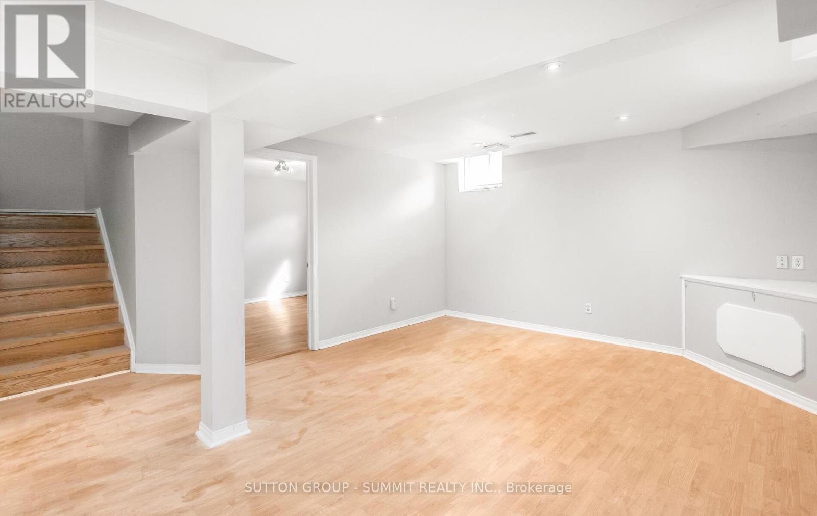 A spacious room with wooden flooring and white walls, featuring a staircase and a fireplace.