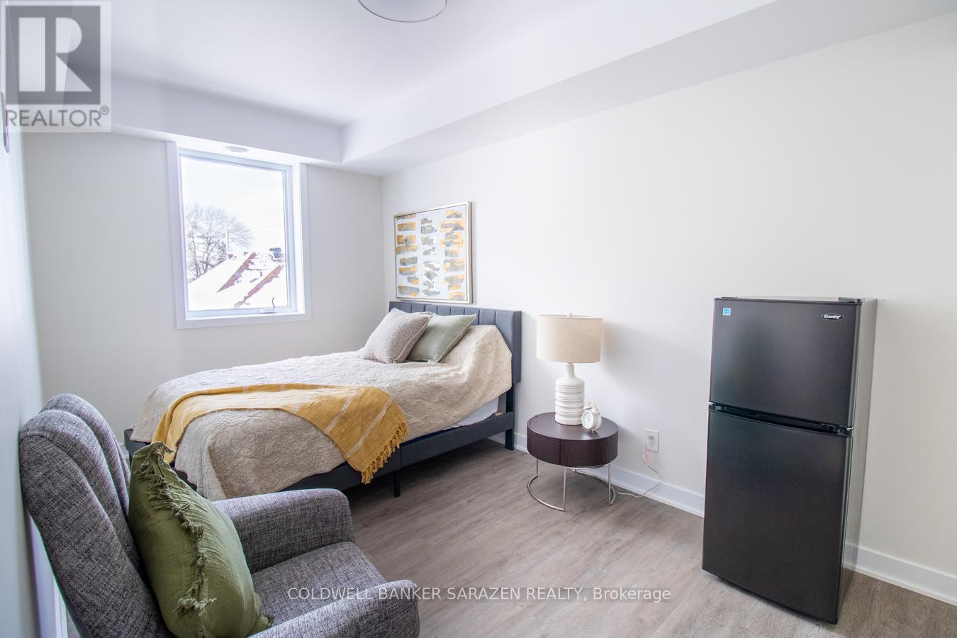 A bedroom with a bed, chair, and a refrigerator.