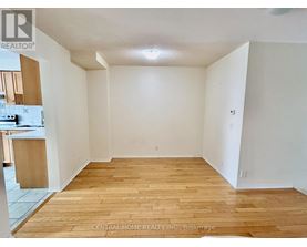 A room with wooden flooring and white walls, with a kitchen area visible in the background.